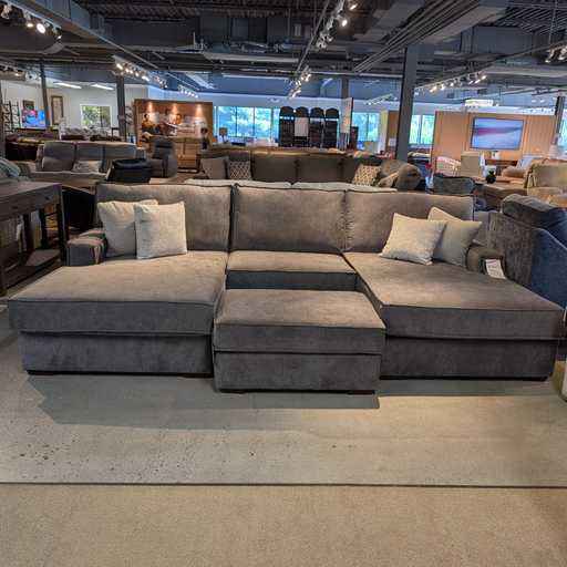 A large gray sectional sofa with a chaise lounge and matching ottoman, styled with several light gray throw pillows in a furniture showroom.