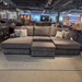 A large gray sectional sofa with a chaise lounge and matching ottoman, styled with several light gray throw pillows in a furniture showroom.