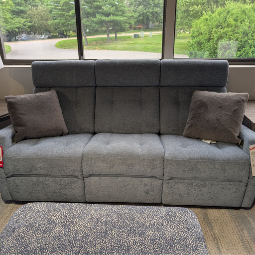 A gray upholstered reclining loveseat sofa with two matching gray throw pillows, positioned in front of large windows showing a green outdoor background.