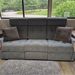 A gray upholstered reclining loveseat sofa with two matching gray throw pillows, positioned in front of large windows showing a green outdoor background.